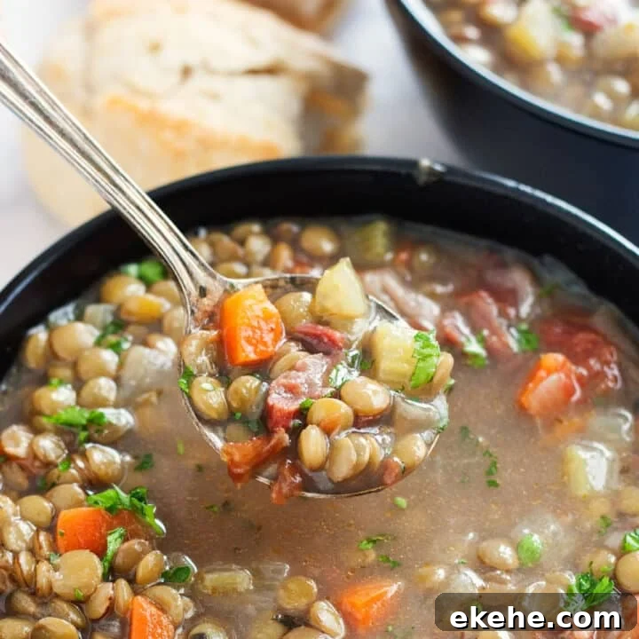 A spoon holding up a scoop of lentil soup with ham from a black bowl of it.