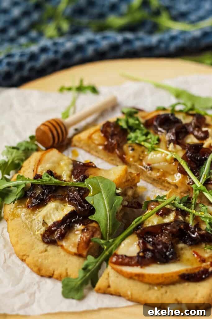 side angle shot of flatbread topped with sliced pears, caramelized onions and brie cheese. Garnished with arugula and honey. Flatbread is on top of parchment on a wooden serving board.