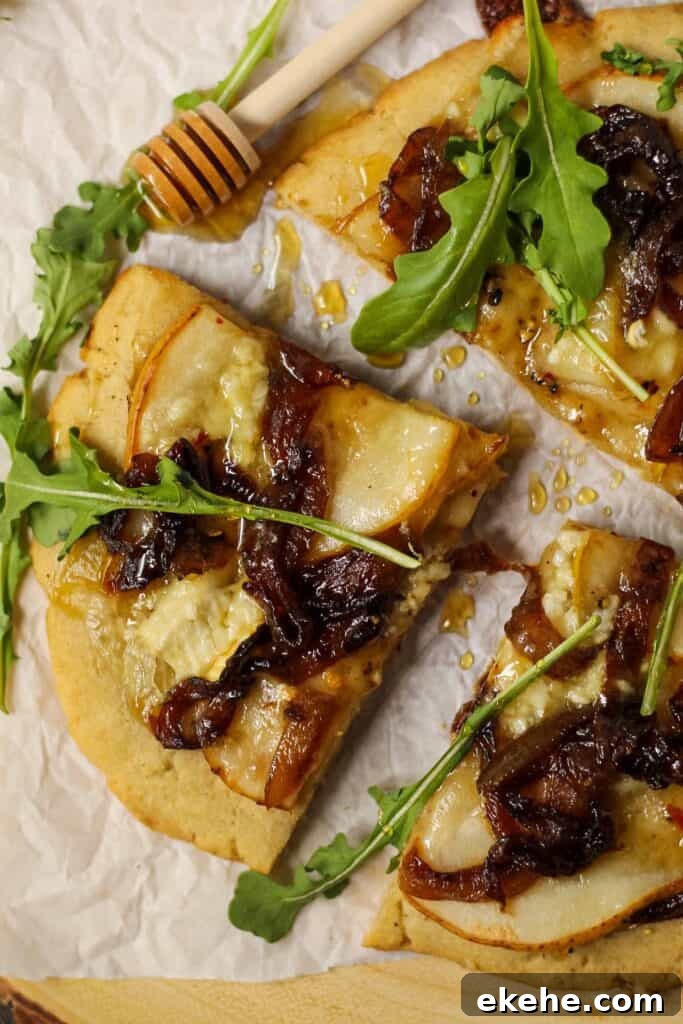 close up overhead shot of flatbread topped with sliced pears, caramelized onions and brie cheese. Garnished with arugula and honey. Flatbread is on top of parchment on a wooden serving board.