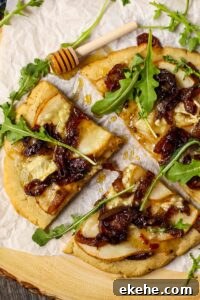 overhead shot of flatbread topped with sliced pears, caramelized onions and brie cheese. Garnished with arugula and honey. Flatbread is on top of parchment on a wooden serving board.
