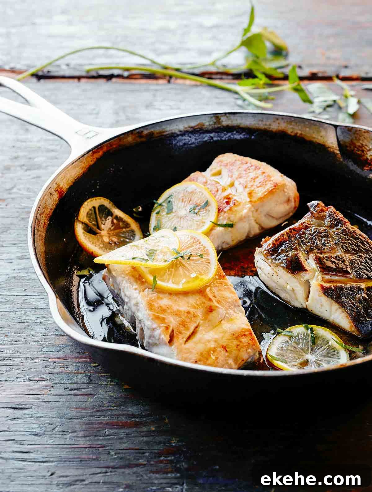 A skillet with three pieces of pan fried fish and lemon slices
