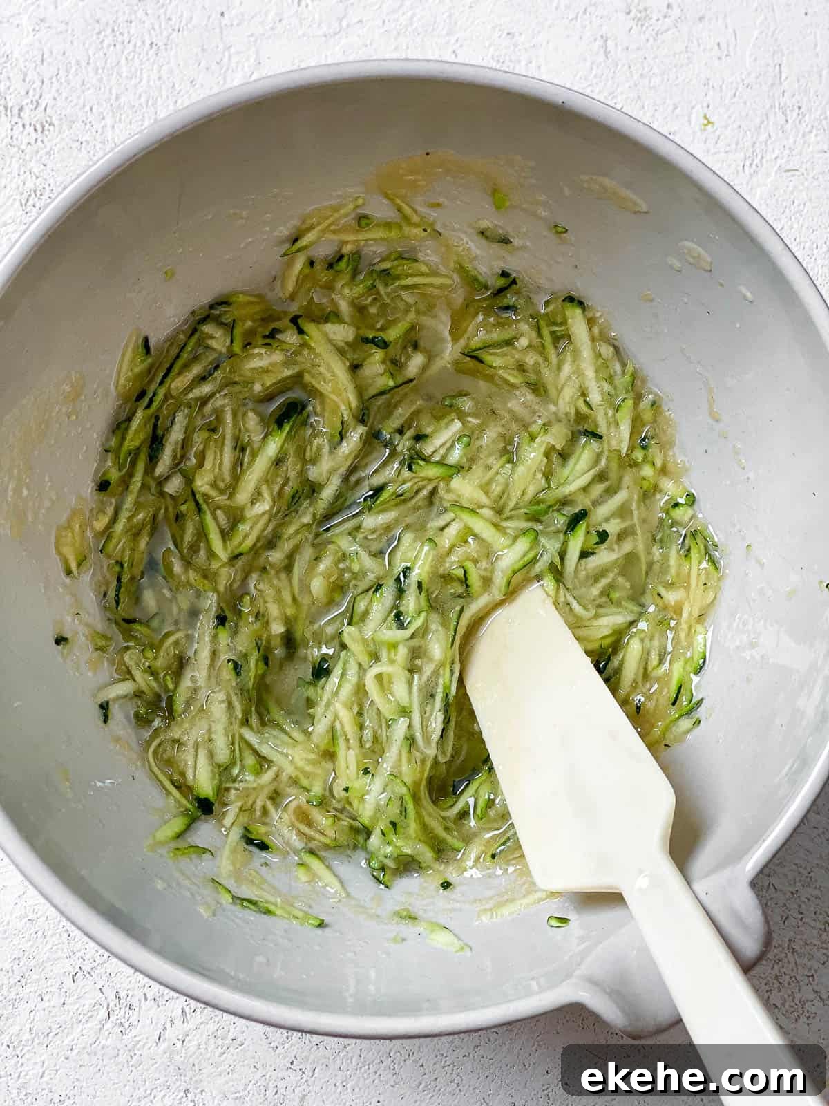 Grated zucchini and wet ingredients in a white bowl with white spatula on a white textured surface.
