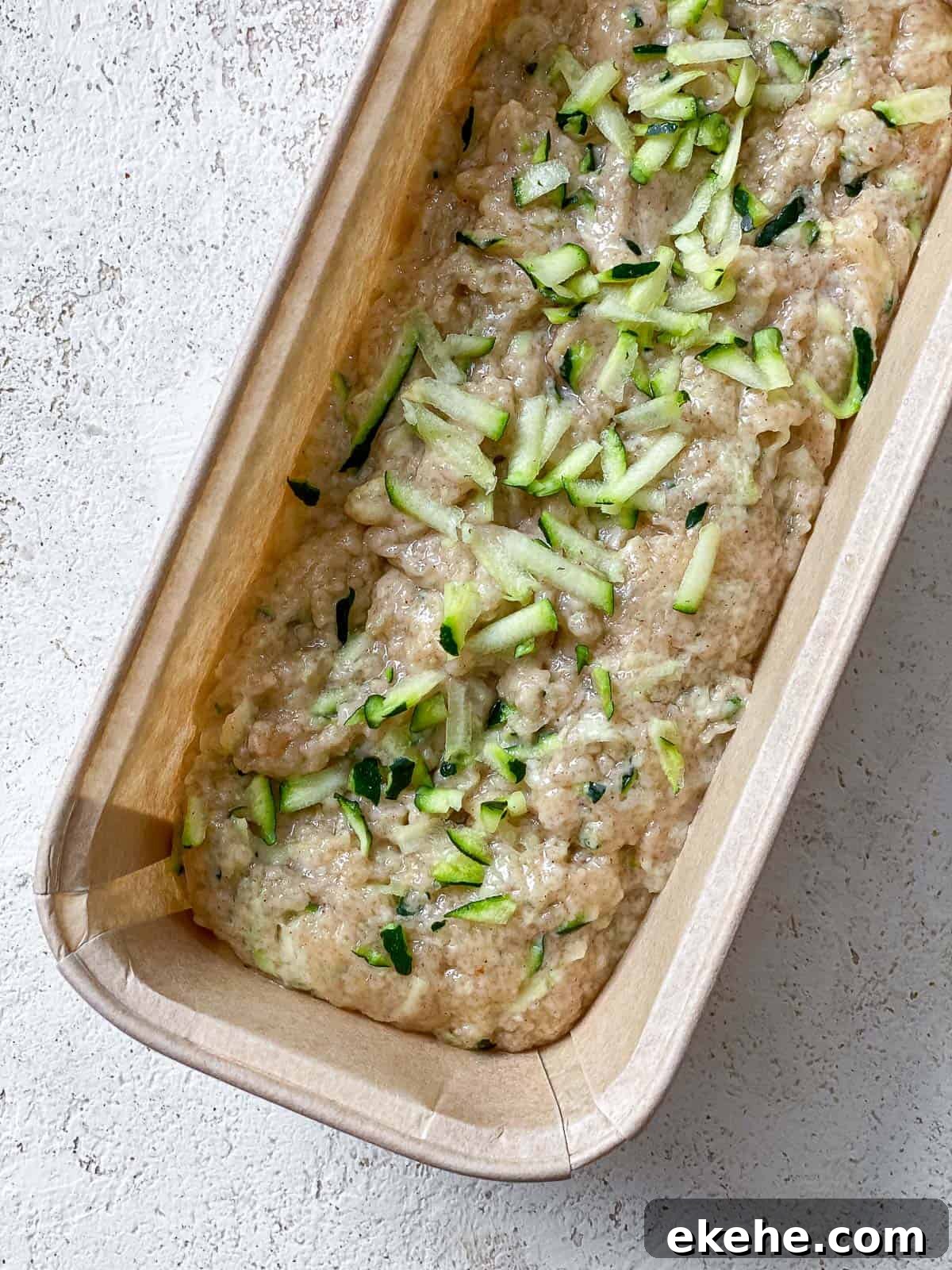 Brown bread loaf pan with zucchini bread batter filled in it sitting on top of a white speckled surface.