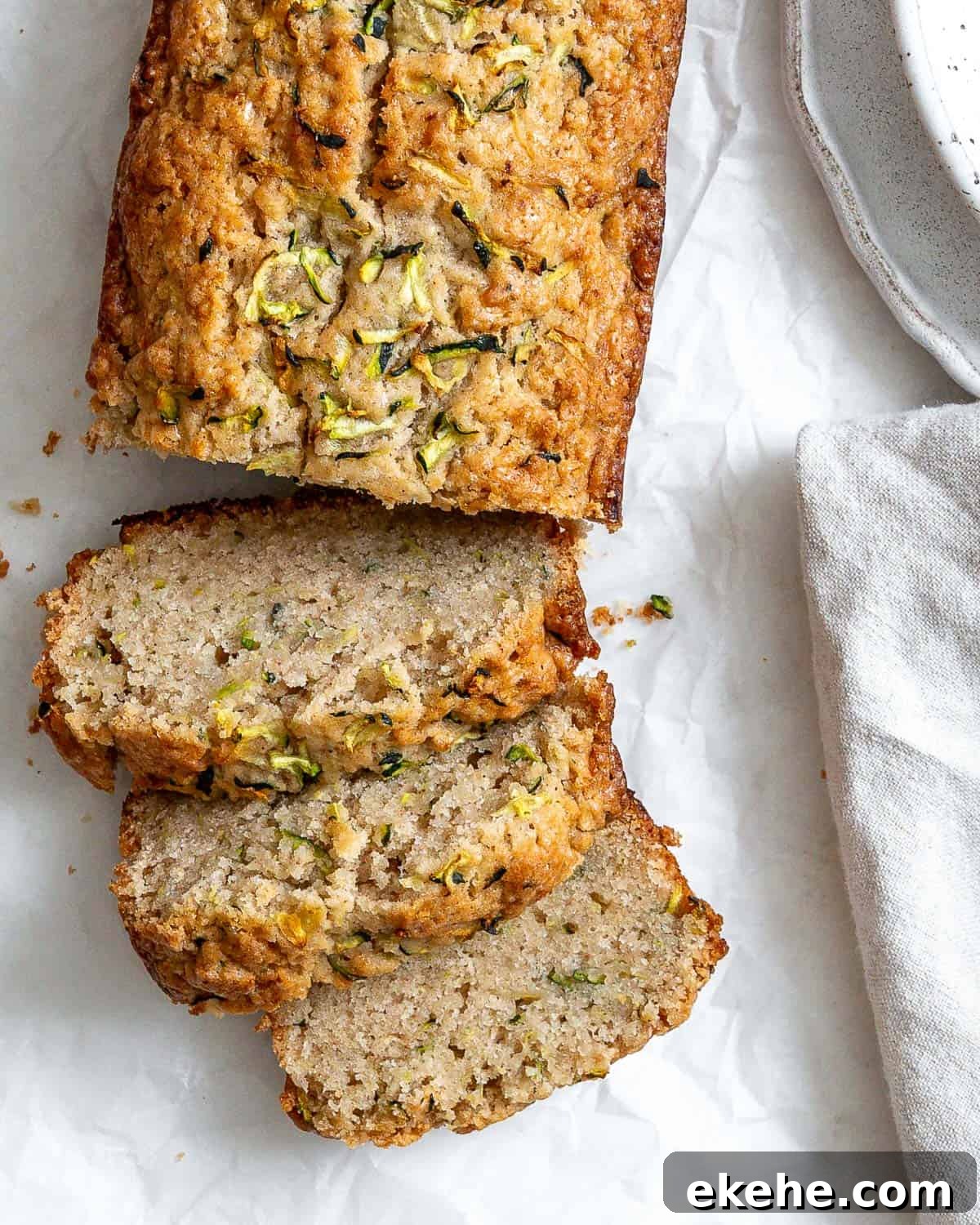 Slices of zucchini bread on white crinkled parchment paper. There's part of a speckled bowl in the top right corner, and part of a gray napkin in the bottom right corner.