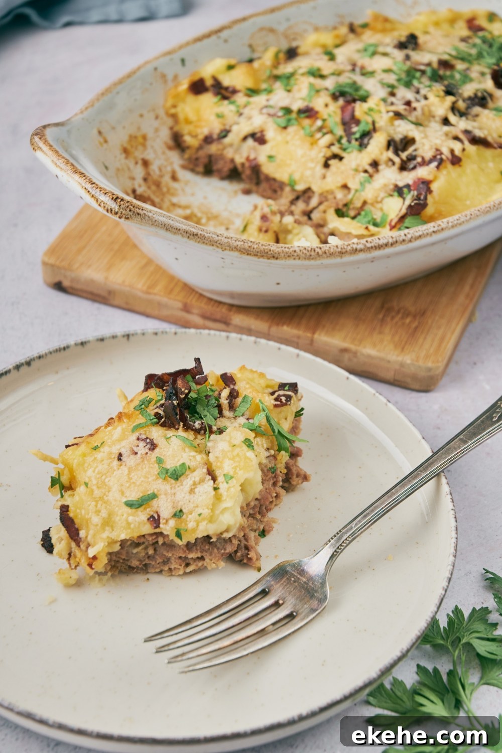 A piece of meatloaf casserole on a plate with a fork and a finished meatloaf casserole in a serving dish behind it.