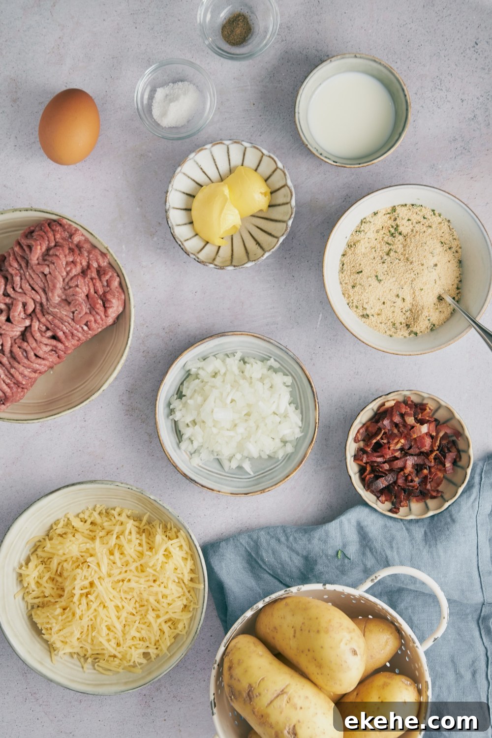Ground meat, shredded cheese, spices, and potatoes in separate bowls on a gray counter.