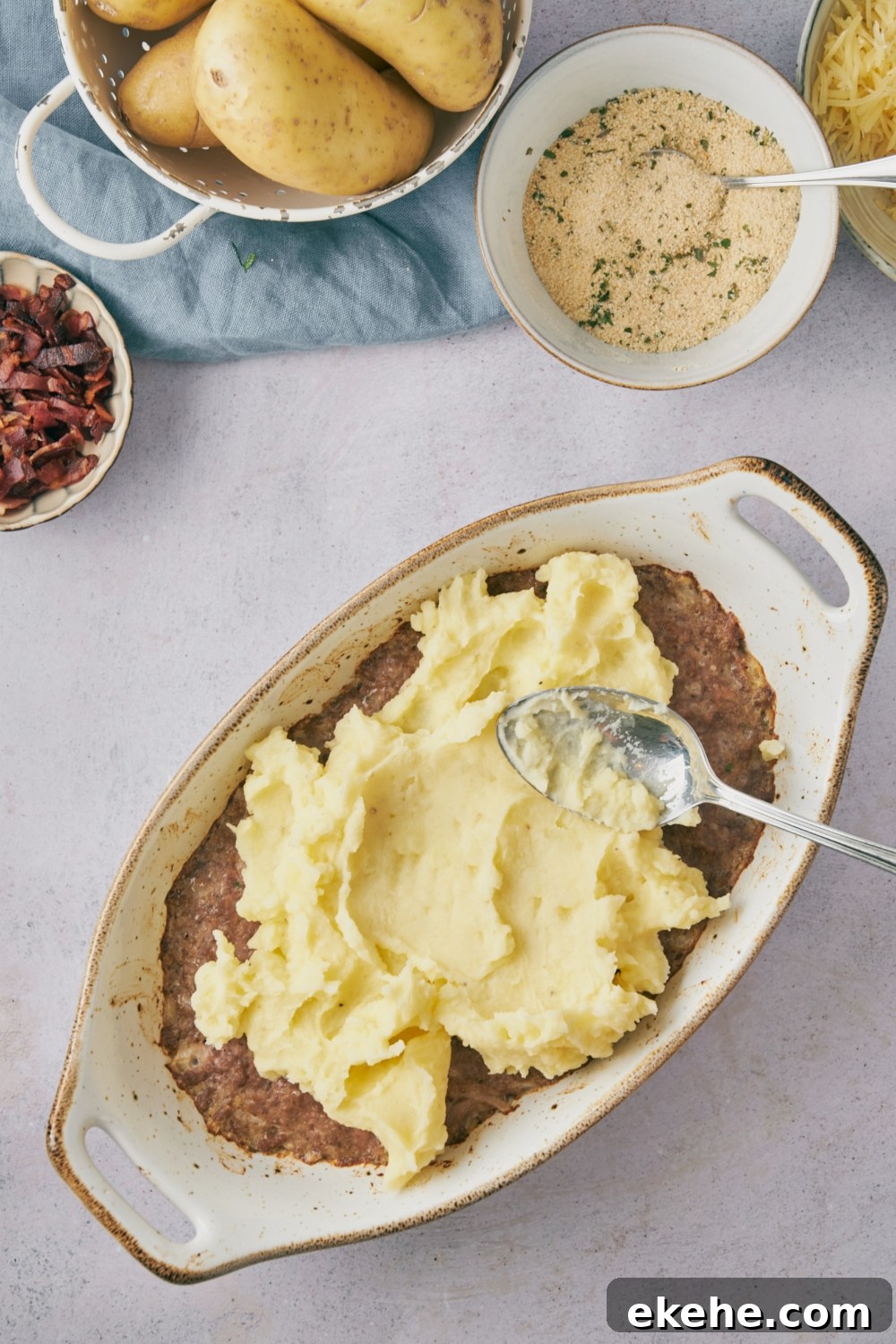 A spoon spreading mashed potatoes over brown meat mixture in a white casserole dish.