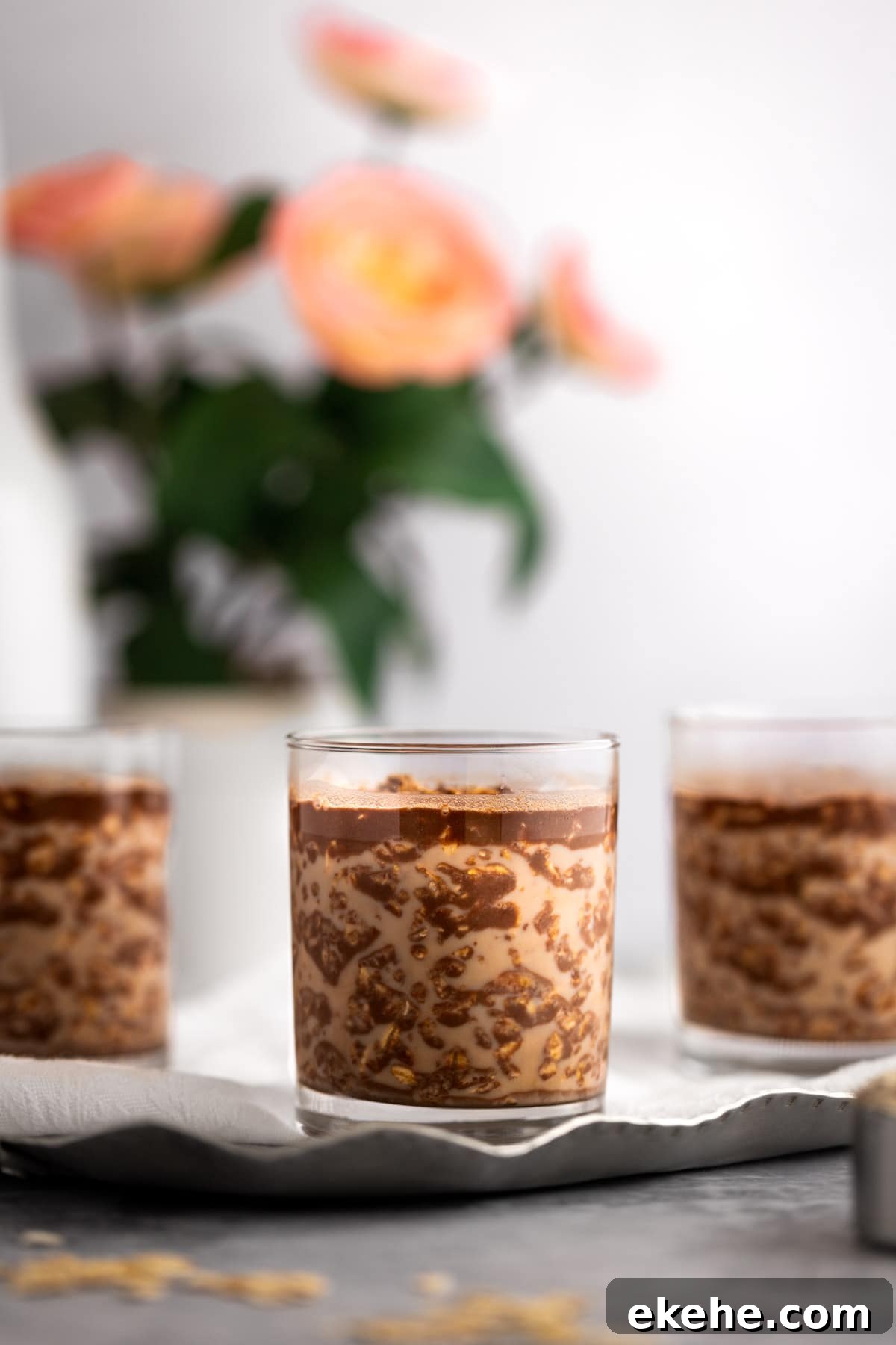 Three glasses of Nutella overnight oats on a metal tray, with pink flowers in the background.