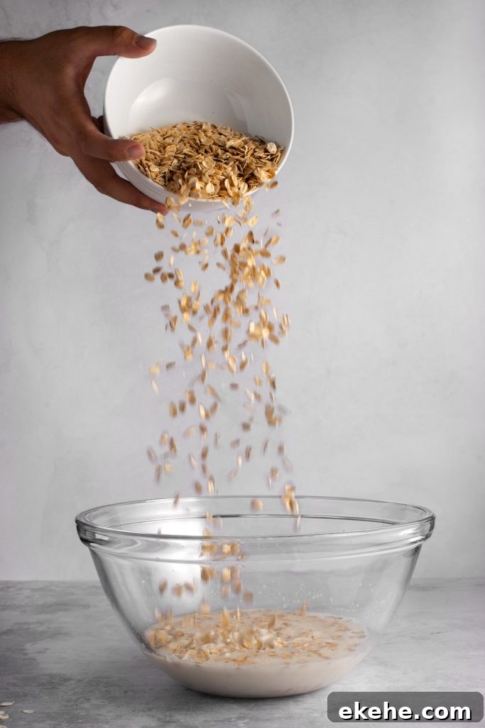 A hand pouring a bowl of overnight oats into a large glass mixing bowl, on a grey background.