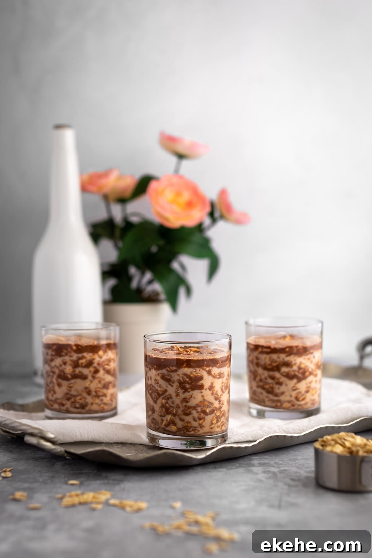Three glasses of overnight oats, on a silver serving platter, with a pot of pink flowers and a white tall bottle in the background.