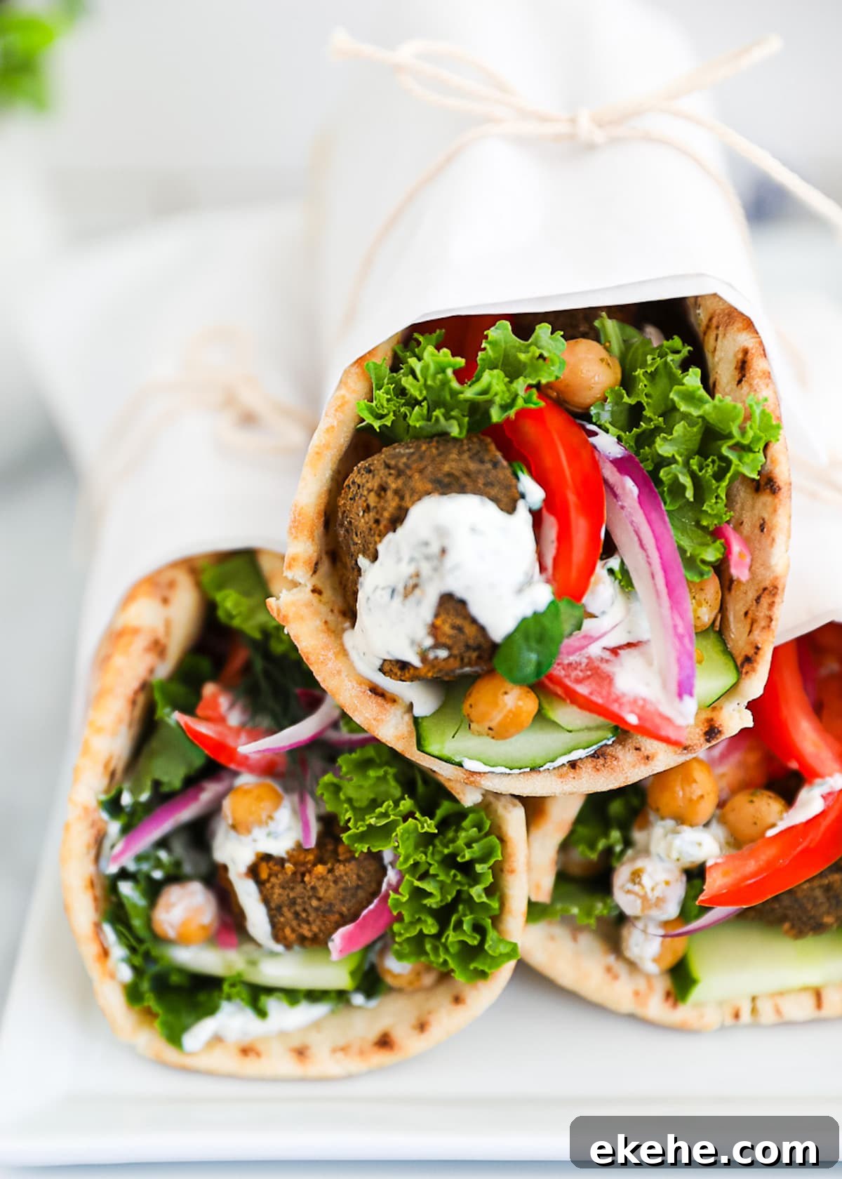 Three vegetarian falafel gyros on a white platter, stuffed with falafel, lettuce, chickpeas, onion, lettuce, tomato, cucumber, and tzatziki sauce.