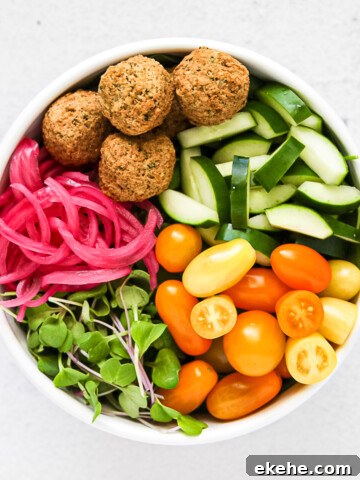 A large white bowl filled will cucumbers, tomatoes, greens, pink pickled red onions, and falafel.