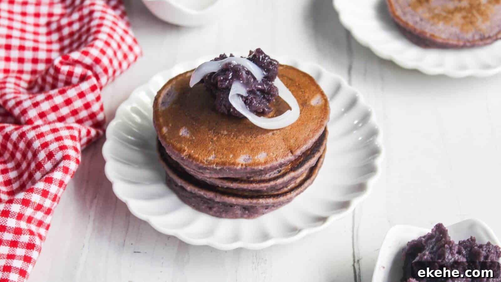 A stack of three purple pancakes topped with sweet purple paste and white strips, served on a white plate with a red checkered cloth nearby.