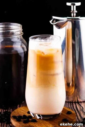 A tall glass of vanilla sweet cream cold brew with a mason jar of cold brew in the background and a french press.