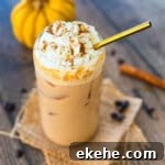 Glass of homemade iced pumpkin spice latte with ice cubes and whipped cream, surrounded by coffee beans and cinnamon sticks.