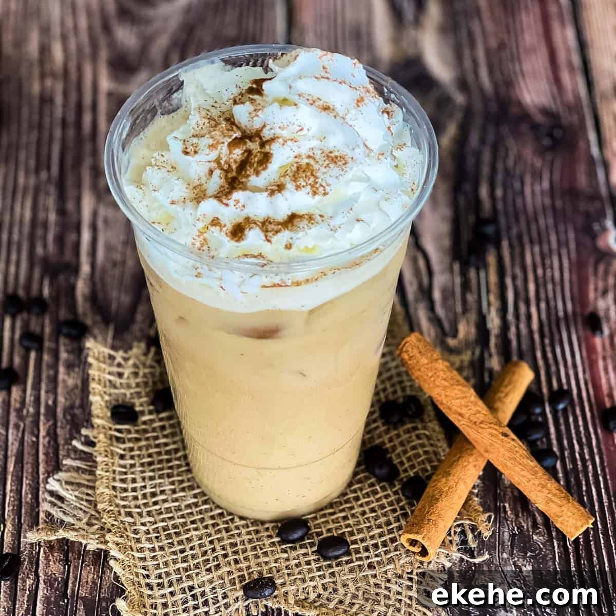 Starbucks copycat iced pumpkin spice latte in a large glass and topped with whip cream and a sprinkle of cinnamon. Two cinnamon sticks beside it with coffee beans.