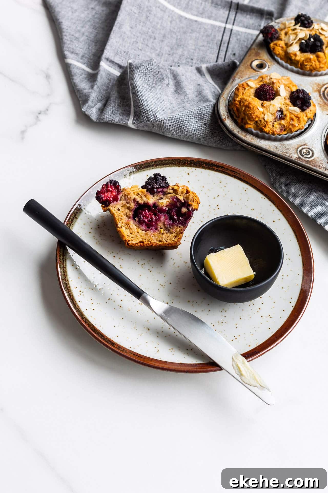 Hearty oat apple muffins with blackberries on a ceramic plate with butter and a knife, vintage muffin pan in background