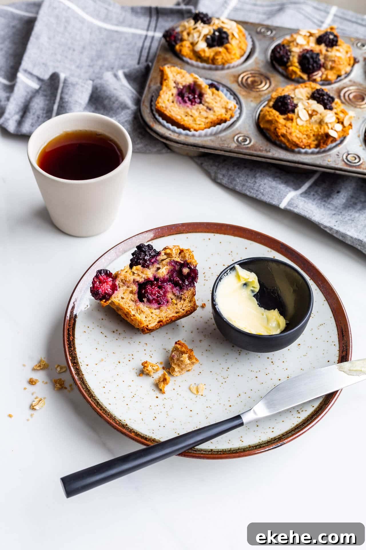 Hearty apple blackberry muffins with oats, one split open on a plate with butter and a knife, served with black tea