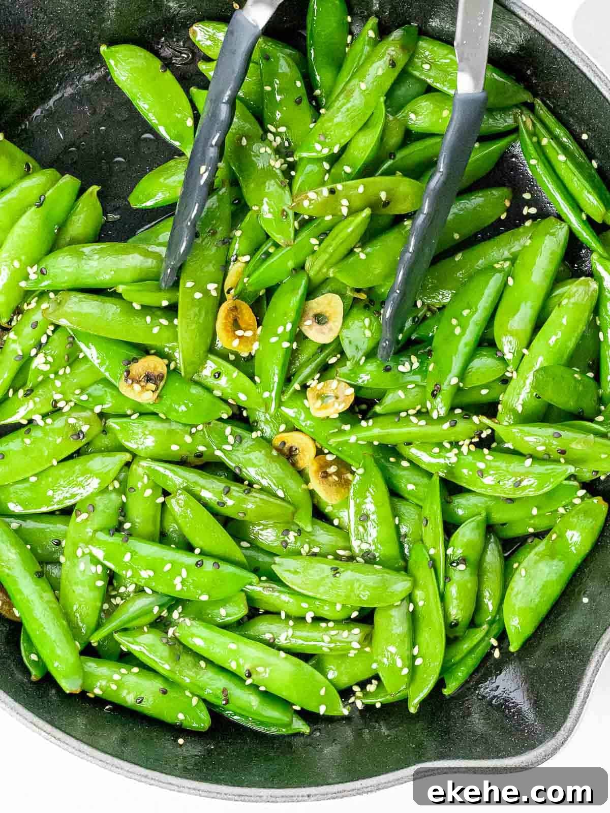 Stir fried sesame sugar snap peas with slices of garlic in a pan with a pair of tongs.