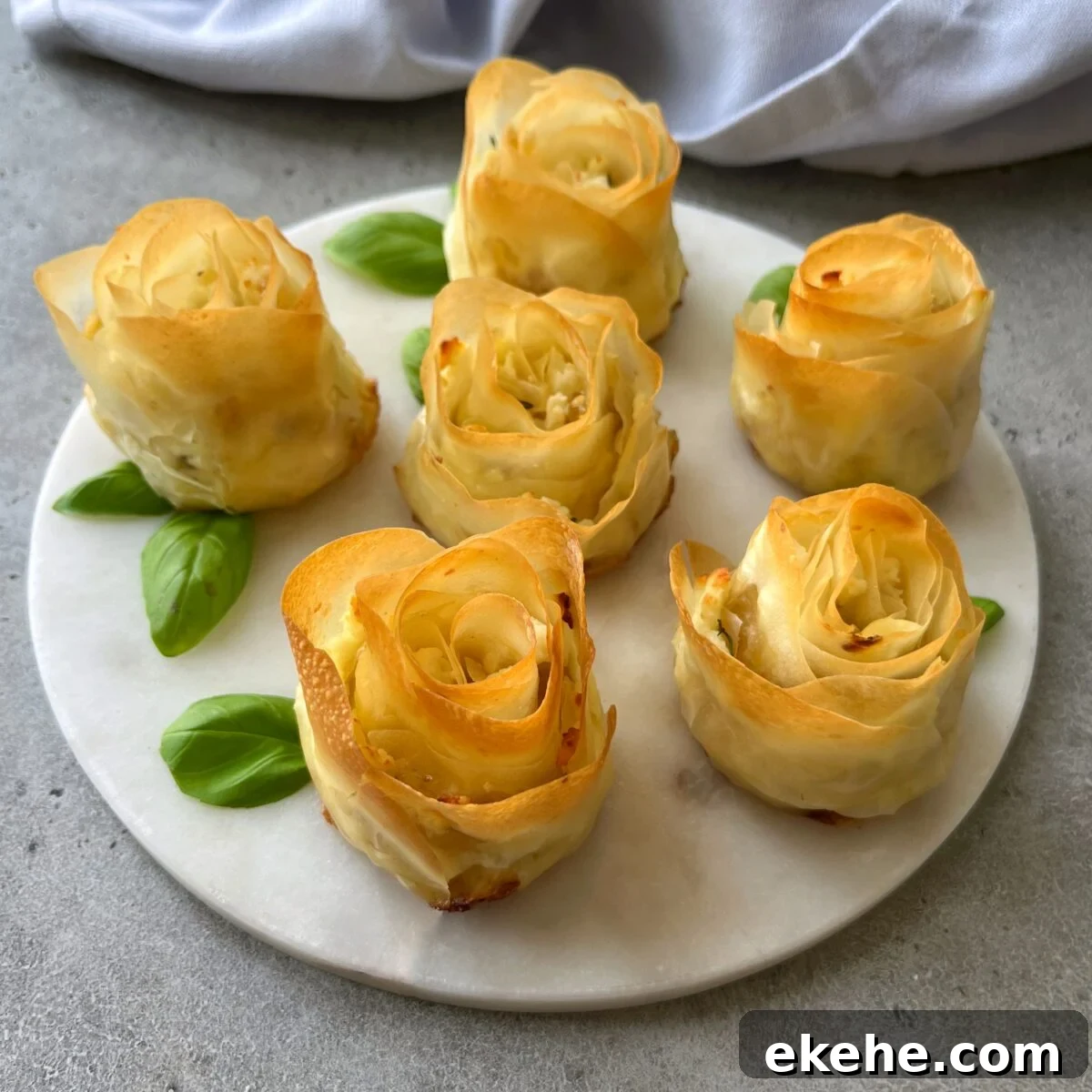 Six Phyllo Appetizers shaped like roses are arranged on a round white plate, each accompanied by green basil leaves, on a gray surface.