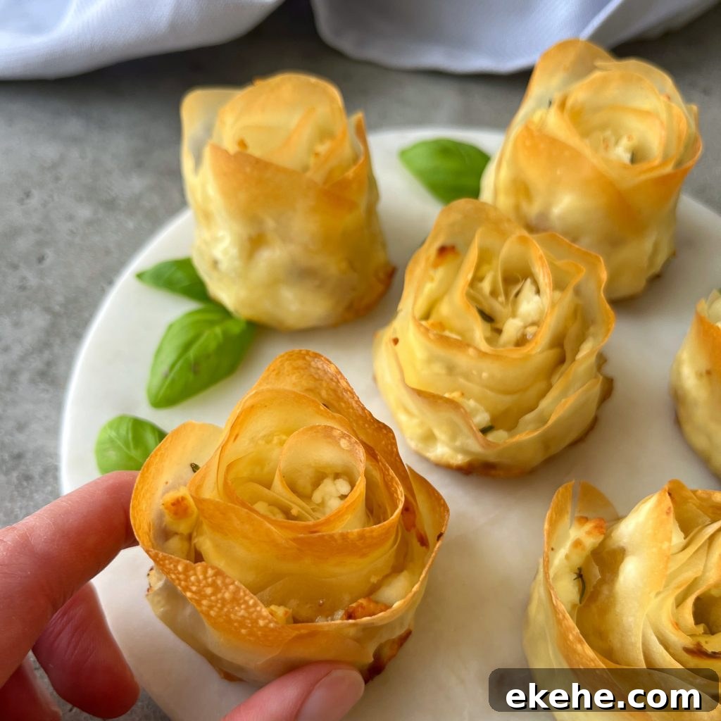 A hand holds a baked pastry shaped like a rose on a white plate, surrounded by more phyllo appetizers and fresh basil leaves. The pastries have golden, crisp edges and a creamy filling.