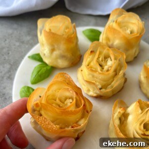 A hand holds a baked pastry shaped like a rose on a white plate, surrounded by more phyllo appetizers and fresh basil leaves. The pastries have golden, crisp edges and a creamy filling.