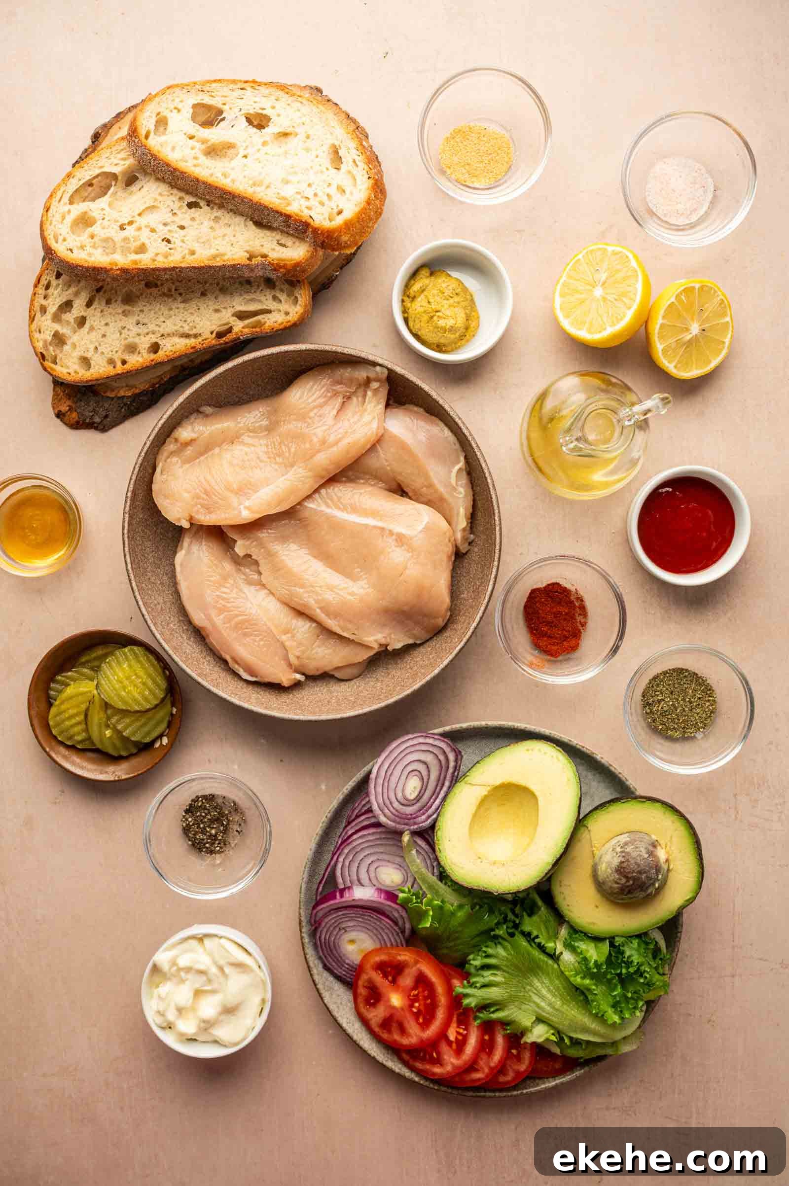 Sliced bread, raw chicken breasts, avocado, tomato, red onion, lettuce, pickles, mayonnaise, spices, mustard, lemon, oil, and sauce arranged on a beige surface.