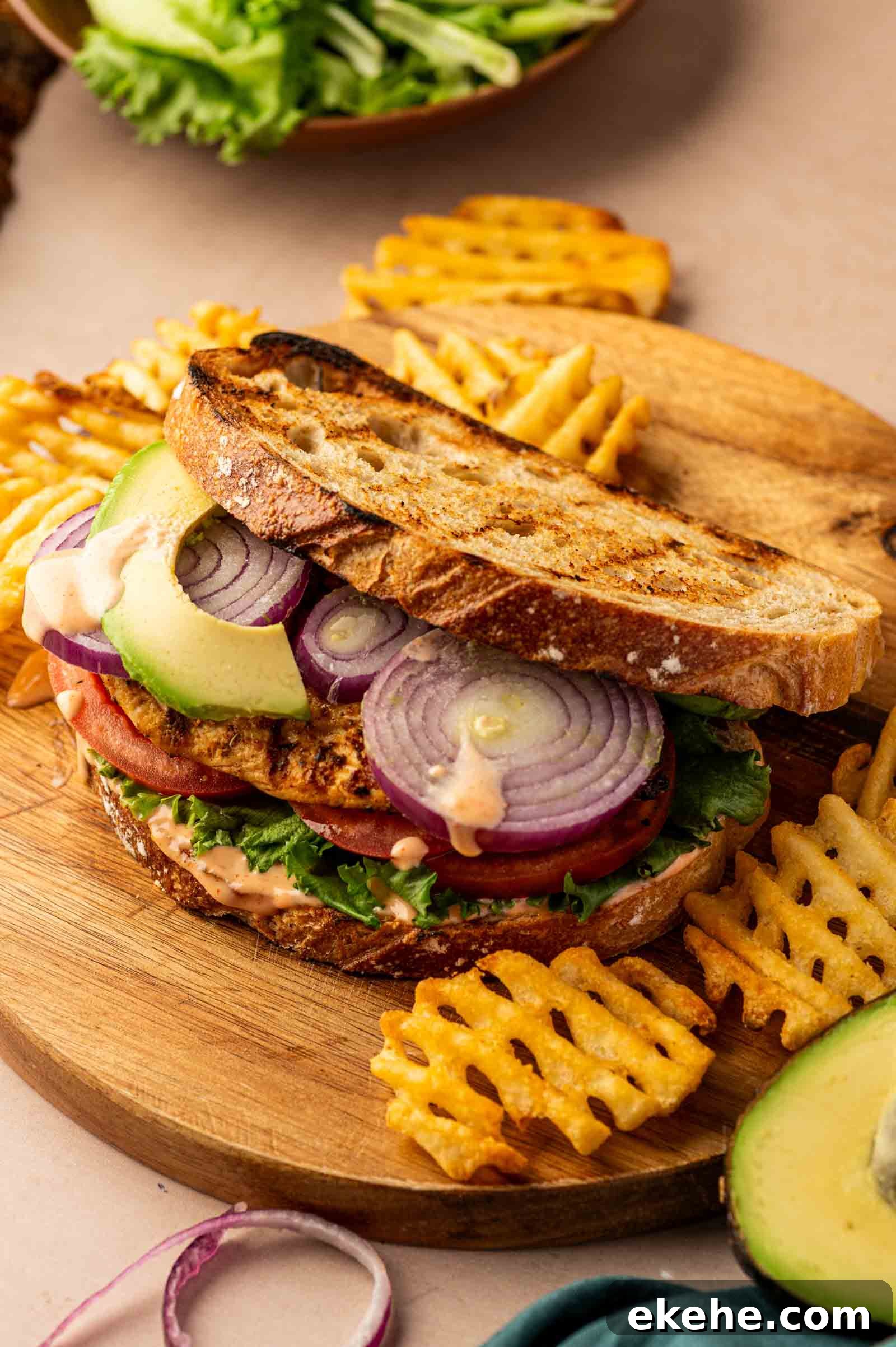 A grilled chicken sandwich with lettuce, tomato, red onion, and avocado on toasted bread, served with waffle fries on a wooden board.