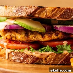 A close-up of a grilled chicken sandwich with lettuce, tomato, red onion, avocado, and sauce on toasted bread, served on a wooden board.