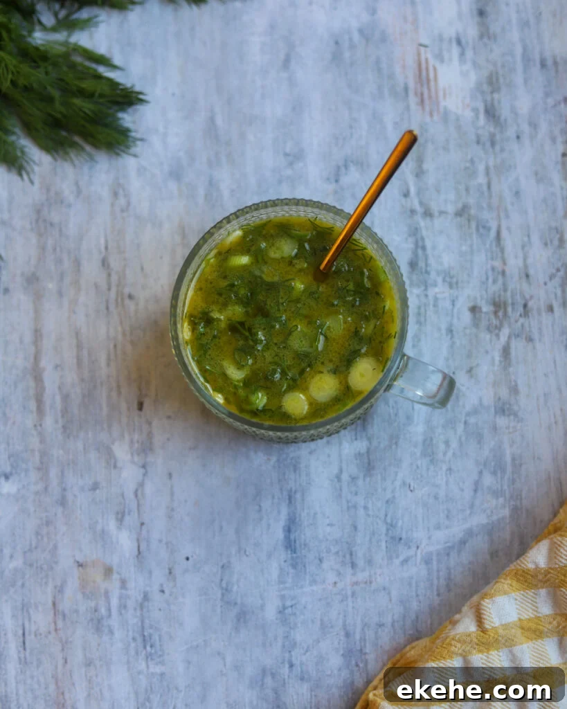 A glass salad bowl is sitting on a gray surface. The bowl is filled with dark green mustard and vinegar salad dressing made with green onions, maple syrup, vinegar and dill. Some fresh dill fronds are showing along with the corner of a checkered cloth
