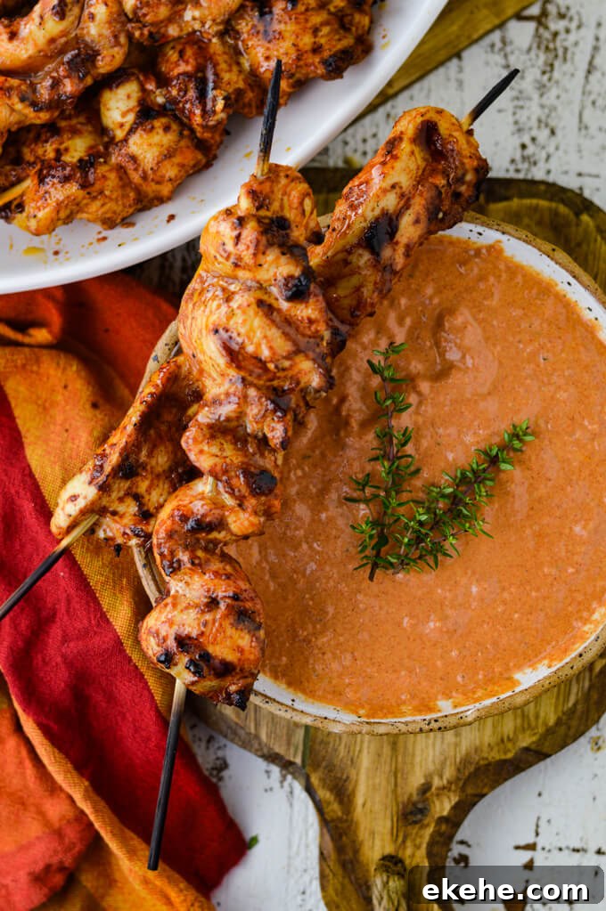 A bowl of Spicy Roasted Tomato and Garlic Aioli sitting on a wooden board. A red & orange napkin sits next to it. Grilled chicken skewers rest on the bowl with more skewers in the top left corner of the photo.