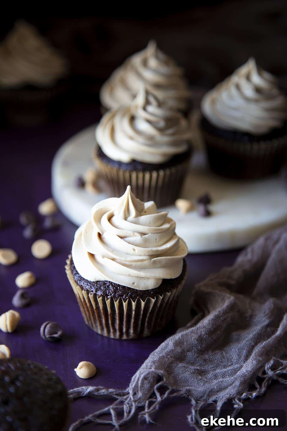 Several chocolate cupcakes with peanut butter frosting on a purple surface with a gray towel
