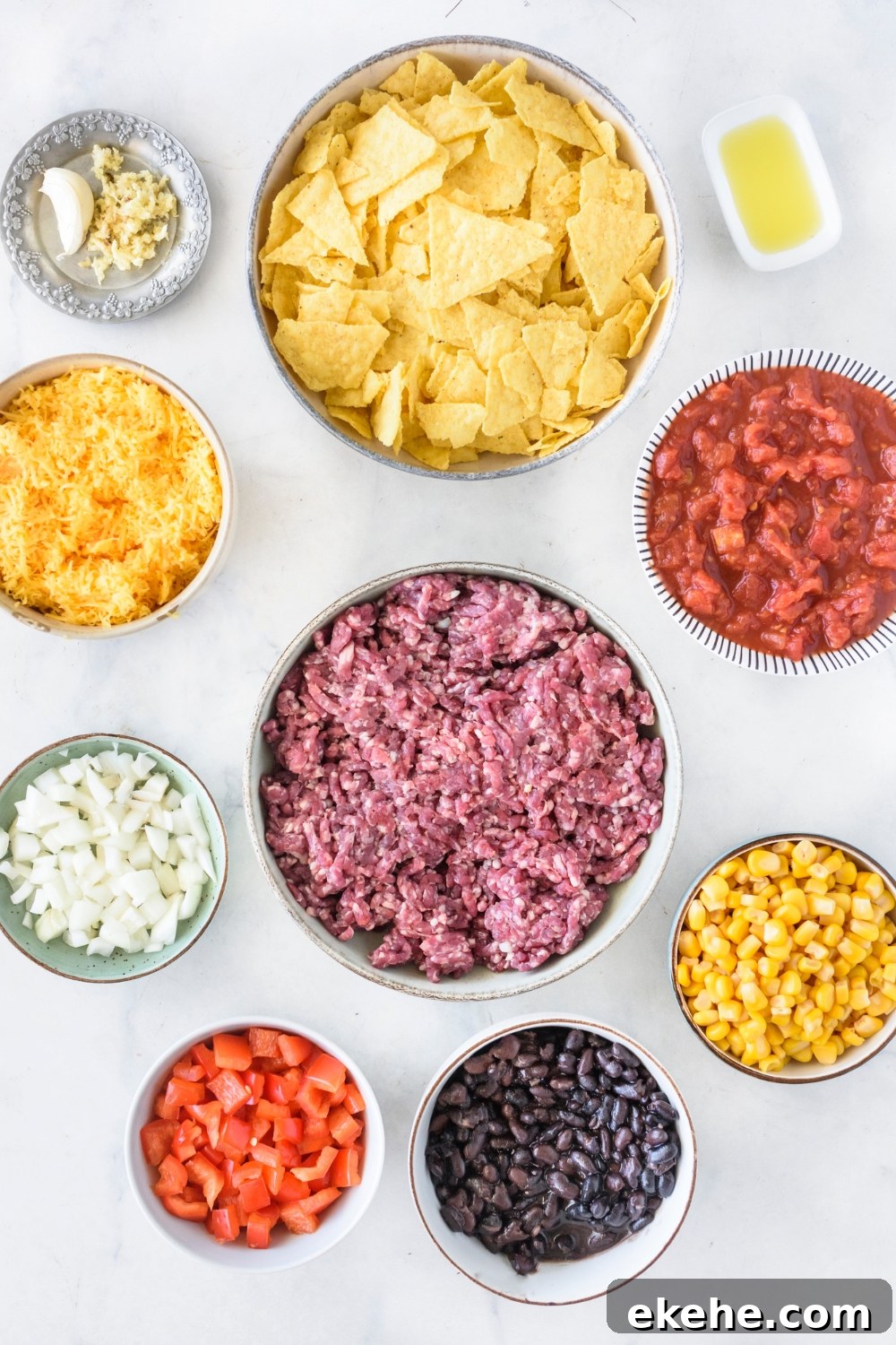 An assortment of ingredients including bowls of crushed tortilla chips, raw ground beef, diced red peppers, diced onion, shredded cheese, salsa, corn, and black beans.