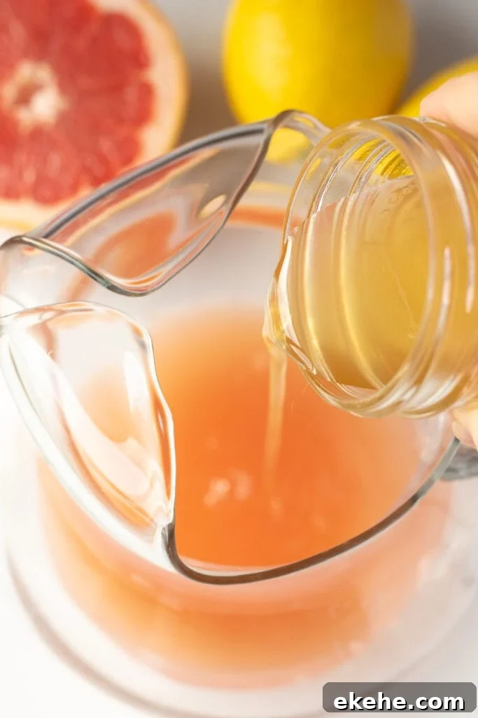 pouring lemon simple syrup into a glass pitcher with grapefruit juice in it.