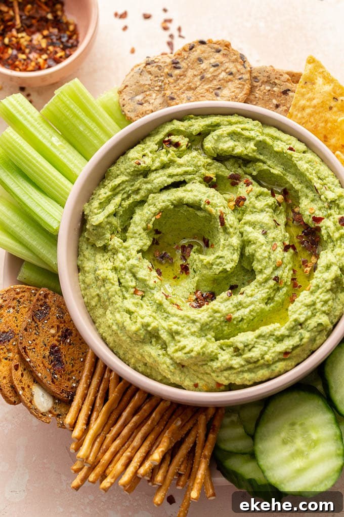 Arugula hummus served in a pink bowl with pretzels, crackers and celery sticks on the side.