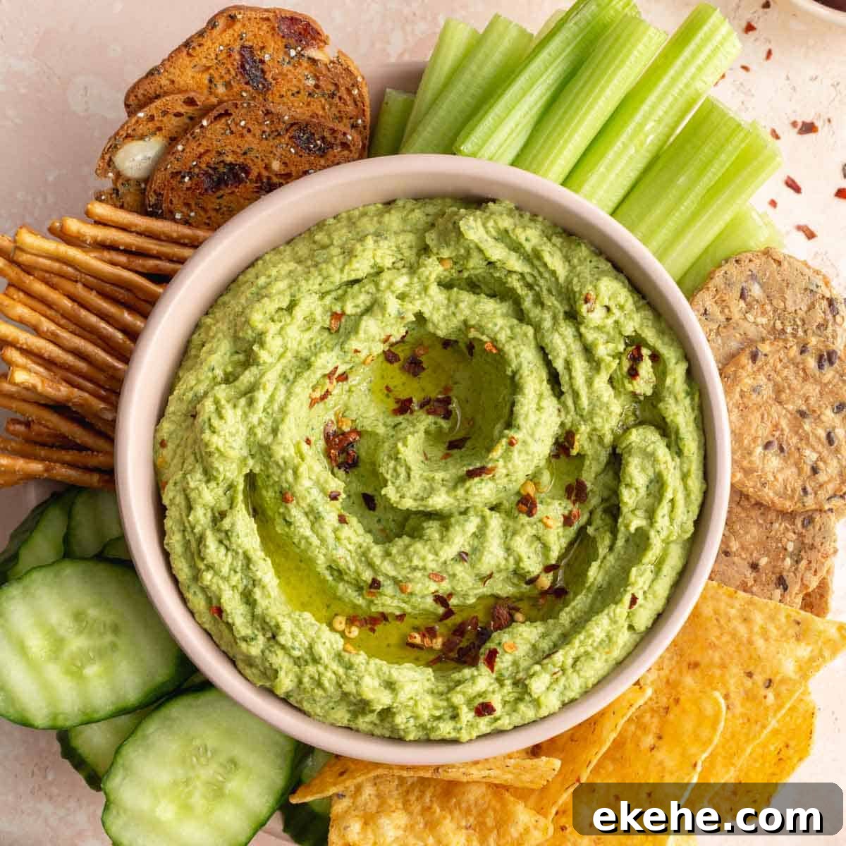 Overhead of arugula hummus in a pink bowl with chips, crackers, pretzels and veggies surrounding it.
